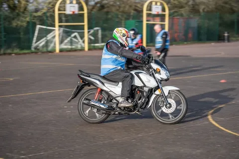 A learner in a bright helmet rides a white motorbike in front of basketball hoops, with an instructor standing behind.