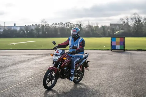 A learner in a grey helmet rides a maroon motorbike towards the camera across wet, sunlit tarmac.