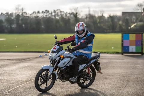 A learner in a white and red helmet rides a white motorbike towards the camera, with a multicoloured target board behind.