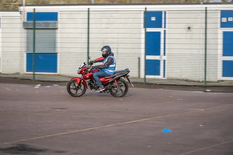 A learner rides a red motorbike along the edge of the practice area in front of a white and blue building.