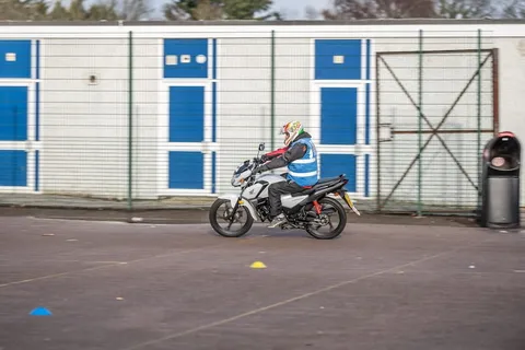 A learner rides a white motorbike past a white building with blue doors and a bin.