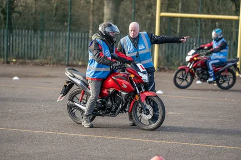 An instructor points ahead from beside a learner on a red motorbike, while another learner rides in the background.
